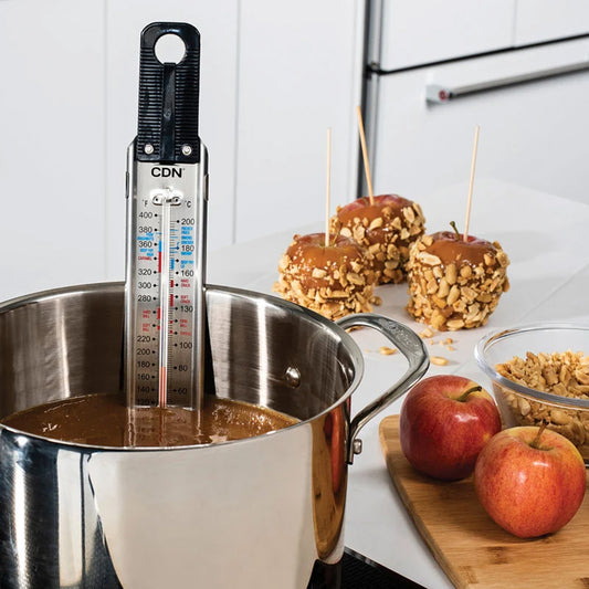 CDN candy thermometer clipped inside a saucepan of caramel with apples and nut-covered caramel apples nearby