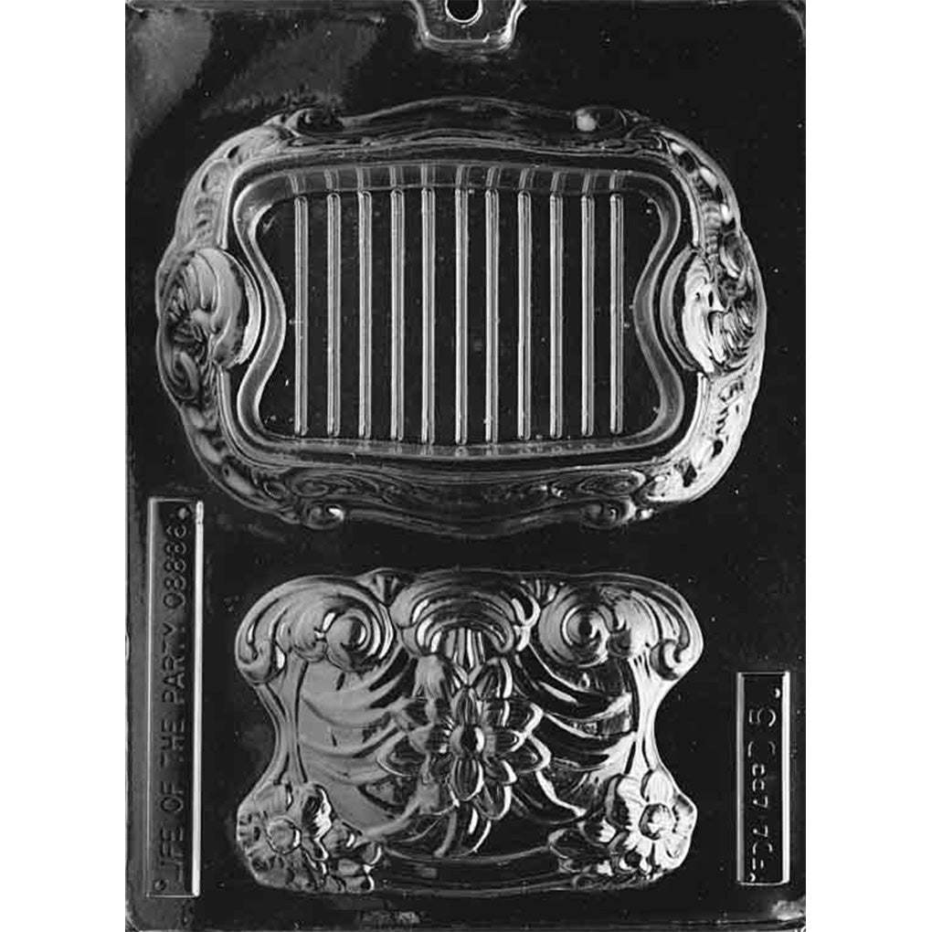 Clear plastic chocolate pour box mold with two large cavities forming a decorative candy dish, featuring ornate scrollwork and a ridged interior panel on a dark background.