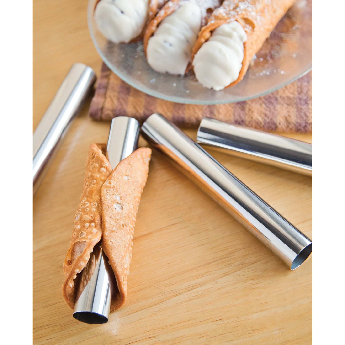 Metal cannoli forms on a wooden surface with a fried cannoli shell wrapped around one tube, with filled cannoli in the background on a serving plate.