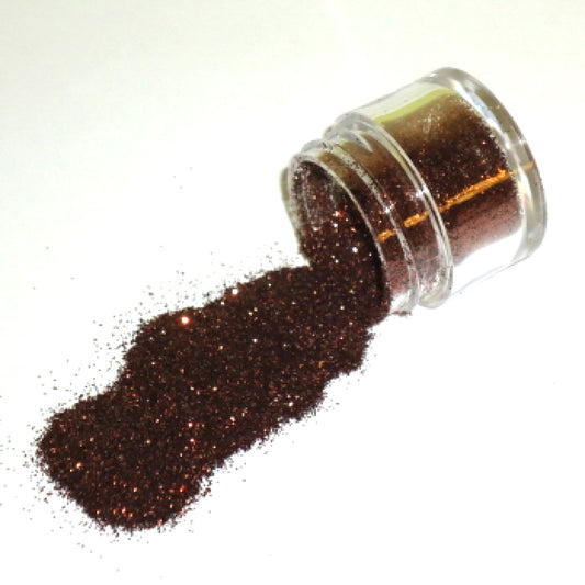 Small clear jar tipped on its side spilling chocolate brown glitter dust onto a white surface, showing fine sparkling brown particles with warm reflective highlights.
