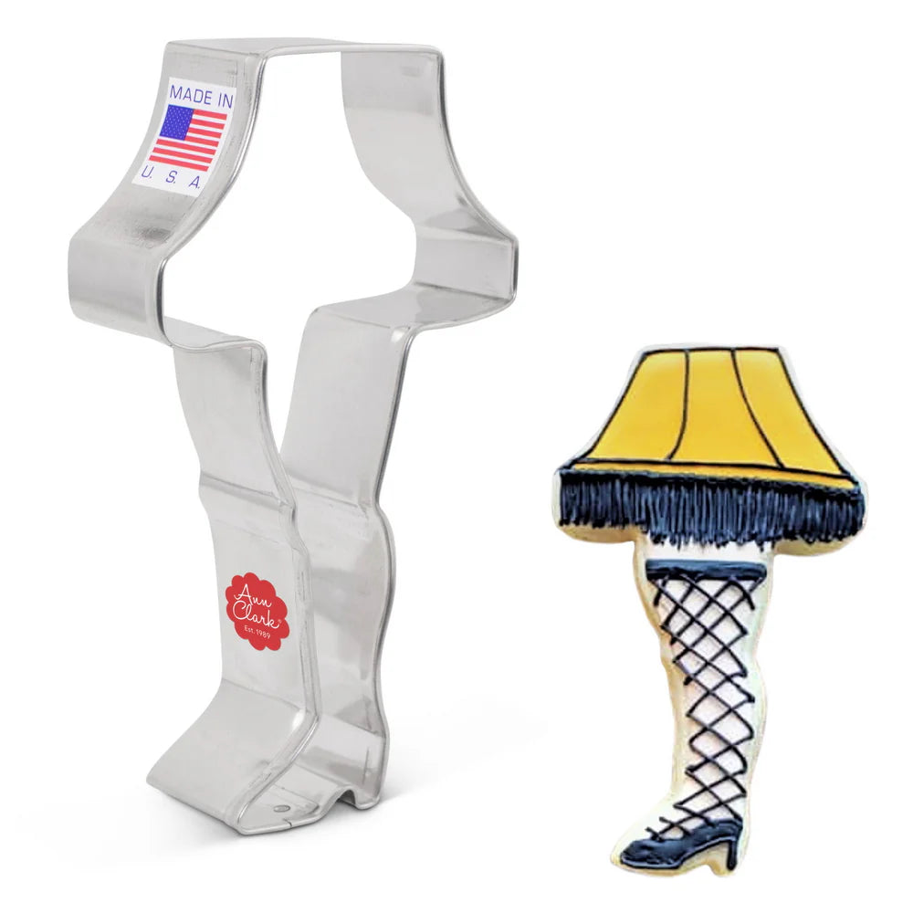 Side view of the leg lamp cookie cutter next to a decorated cookie featuring a yellow lampshade with black fringe and a leg with fishnet stockings and a high heel.