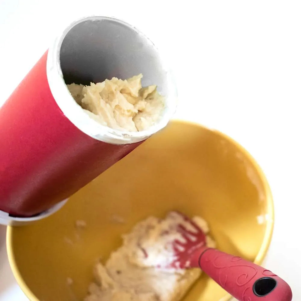 Churro dough being loaded into the churro maker above a mixing bowl with a red spatula.