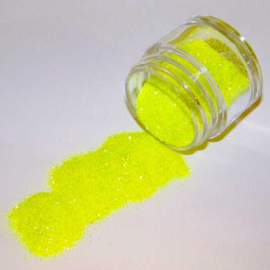 Small clear jar tipped on its side spilling bright citrus yellow-green glitter dust onto a white surface, with fine sparkling particles visible throughout.