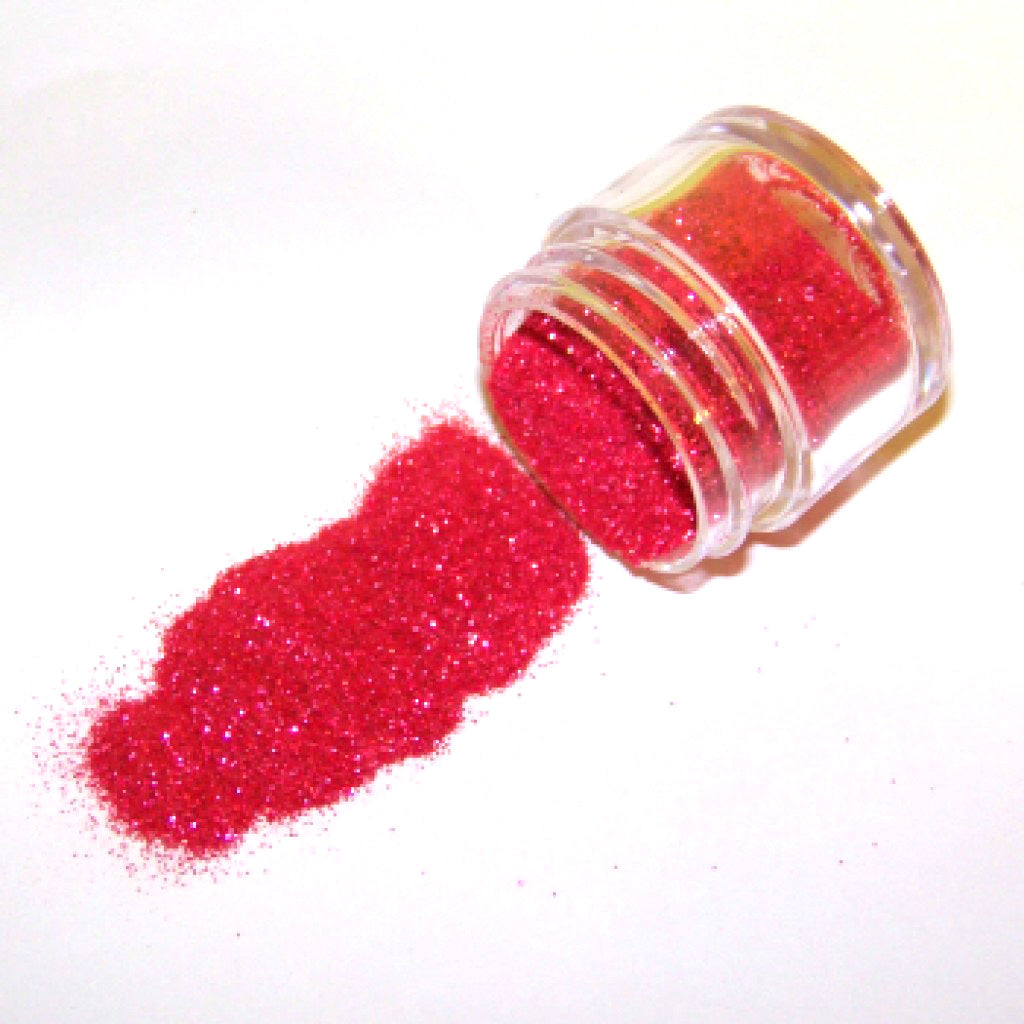 Small clear jar tipped on its side spilling bright crimson red glitter dust onto a white surface, with fine sparkling particles visible throughout.