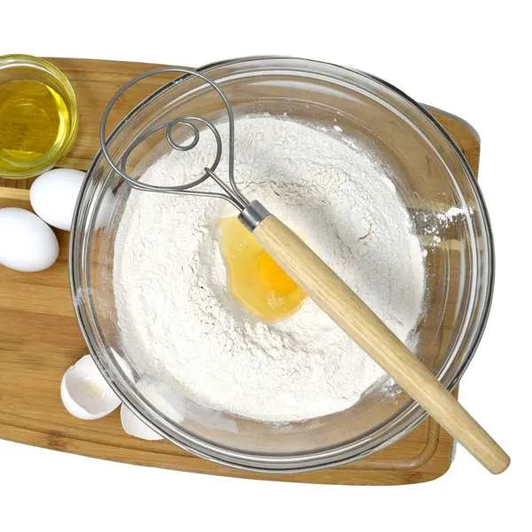 A Danish dough whisk with a wooden handle and metal loop mixes flour and an egg in a glass bowl on a wooden board, surrounded by other baking ingredients.