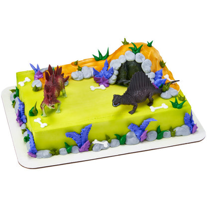 Sheet cake decorated with dinosaur toppers, foliage, and a rocky cave design featuring a Stegosaurus and Spinosaurus figurine.