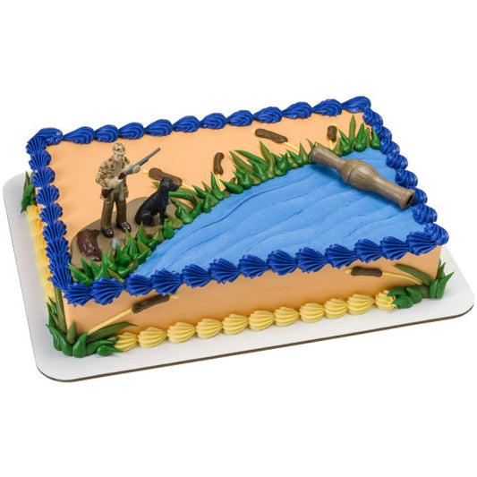 Rectangular cake decorated with duck hunting scene featuring a hunter, dog, duck call, and blue icing pond.
