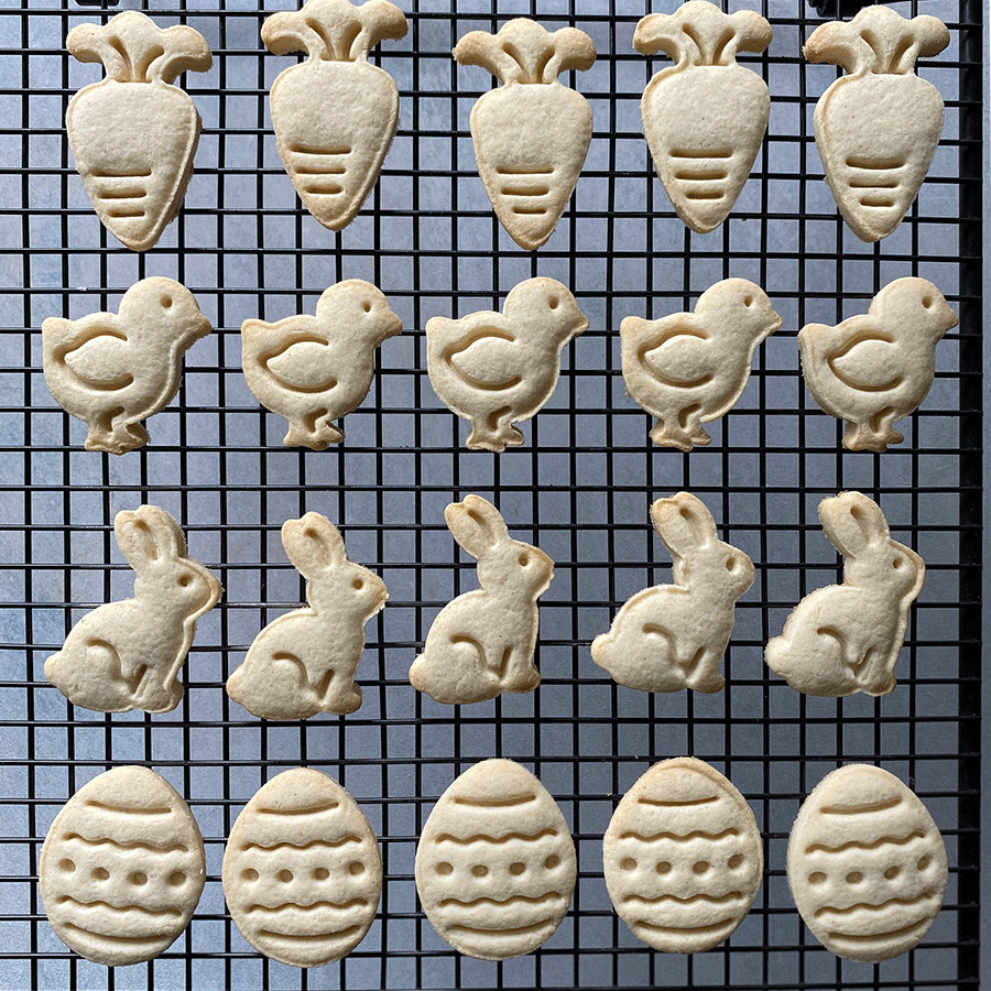 A cooling rack filled with freshly baked cookies featuring stamped Easter designs, including carrots, bunnies, chicks, and eggs.