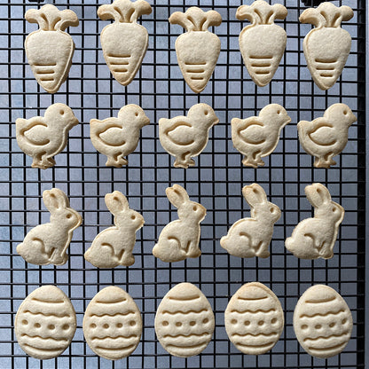 A cooling rack filled with freshly baked cookies featuring stamped Easter designs, including carrots, bunnies, chicks, and eggs.