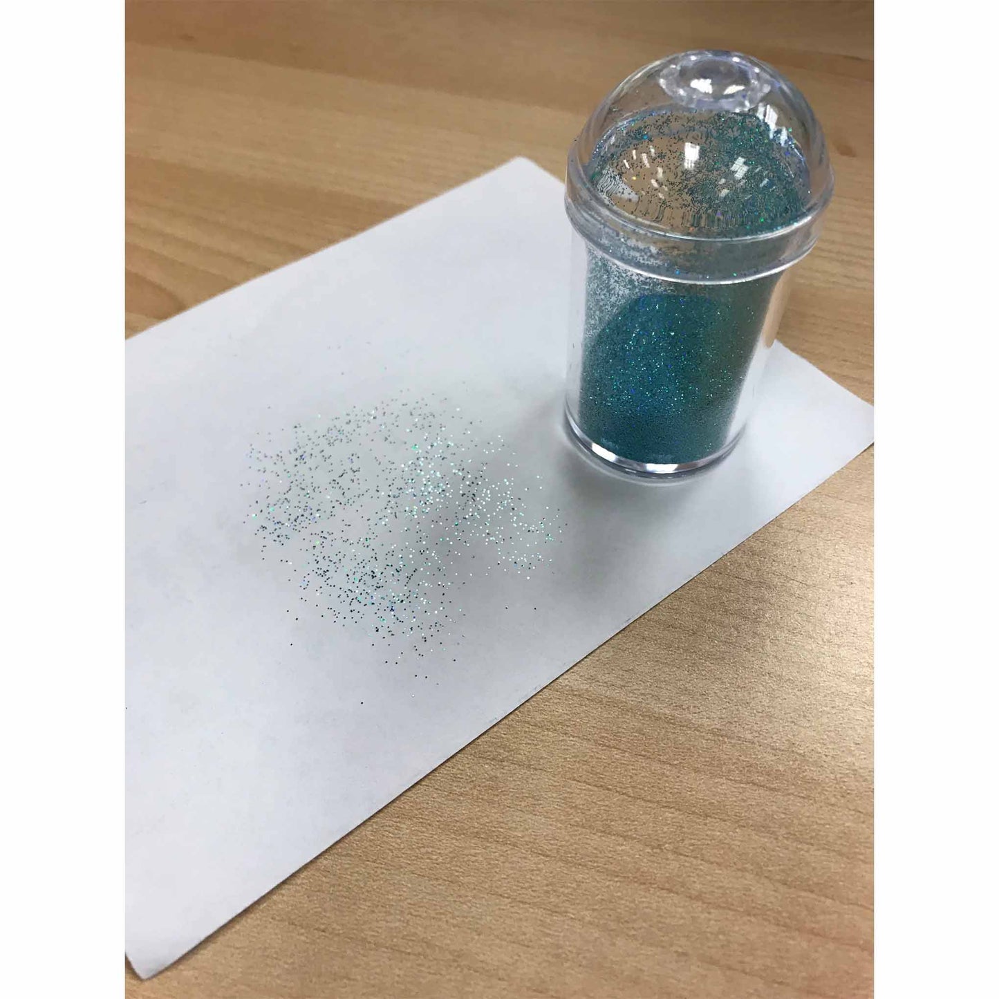 Clear domed-lid shaker standing on a sheet of white paper on a wood table, with scattered fine blue-green edible glitter sprinkled in a soft gradient nearby.