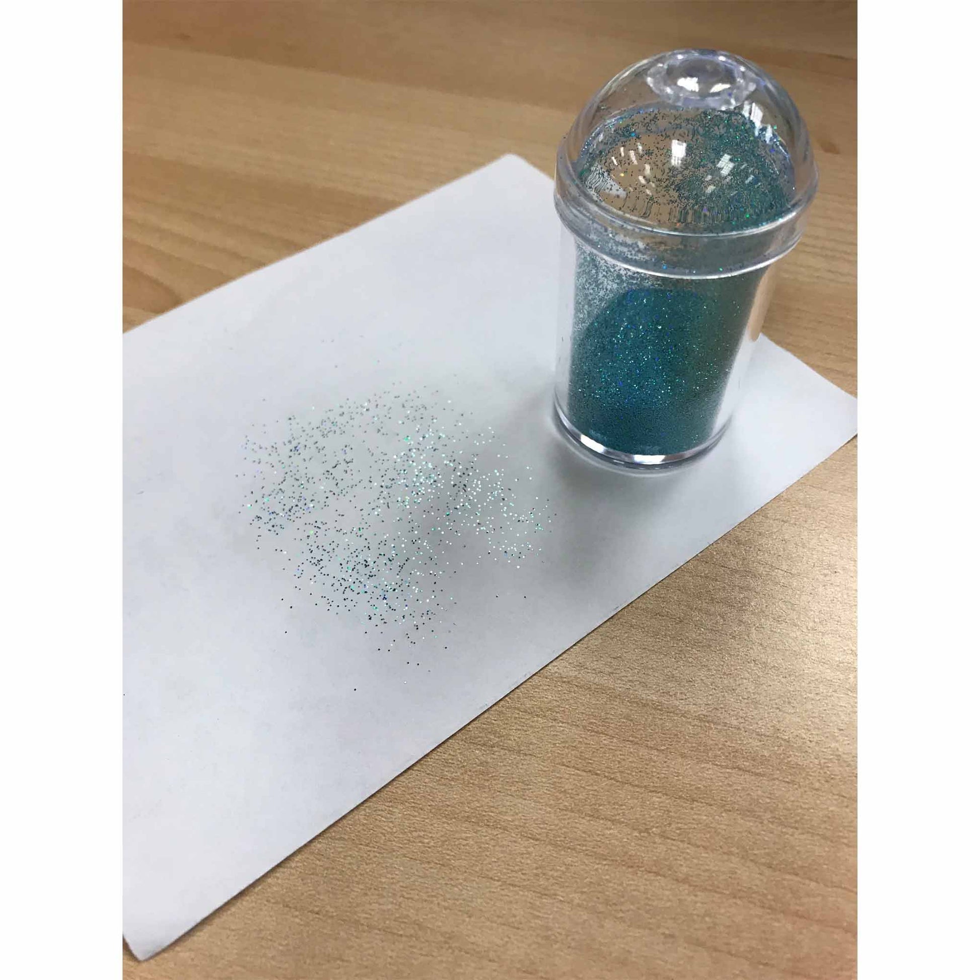 Clear domed-lid shaker standing on a sheet of white paper on a wood table, with scattered fine blue-green edible glitter sprinkled in a soft gradient nearby.