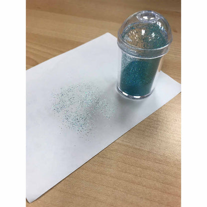 Clear domed-lid shaker standing on a sheet of white paper on a wood table, with scattered fine blue-green edible glitter sprinkled in a soft gradient nearby.