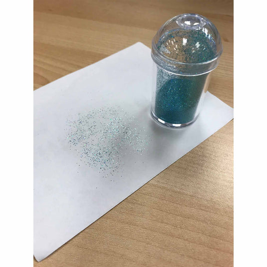 Clear domed-lid shaker standing on a sheet of white paper on a wood table, with scattered fine blue-green edible glitter sprinkled in a soft gradient nearby.