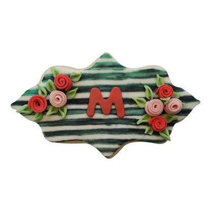 Decorated cookie shaped like an elongated plaque with a green striped background, red fondant "M," and floral details.