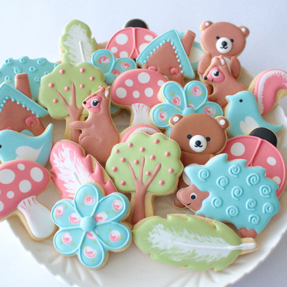 A platter of pastel-colored woodland-themed cookies, including bears, trees, mushrooms, birds, and feathers with intricate icing designs.