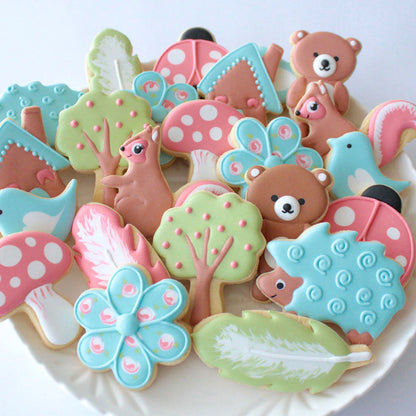 A platter of pastel-colored woodland-themed cookies, including bears, trees, mushrooms, birds, and feathers with intricate icing designs.