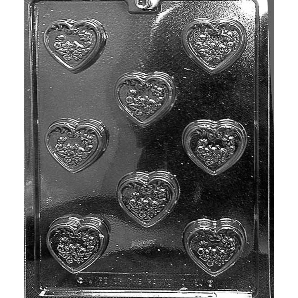 Clear plastic candy mold photographed on a black background with seven heart-shaped cavities, each featuring a raised scroll border and a textured floral leaf pattern inside the heart.