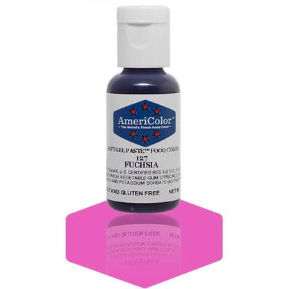 A bottle of AmeriColor Fuchsia soft gel paste food coloring with a white cap and blue label adorned with red stars, standing on a vibrant pink reflective surface with the bottle’s reflection visible.