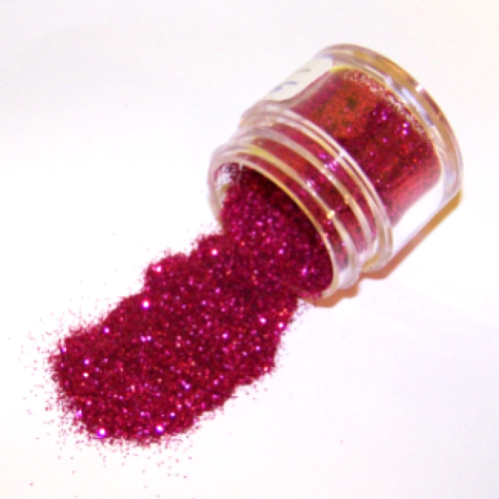 Small clear jar tipped on its side spilling bright fuchsia glitter dust onto a white surface, with fine sparkling particles visible throughout.
