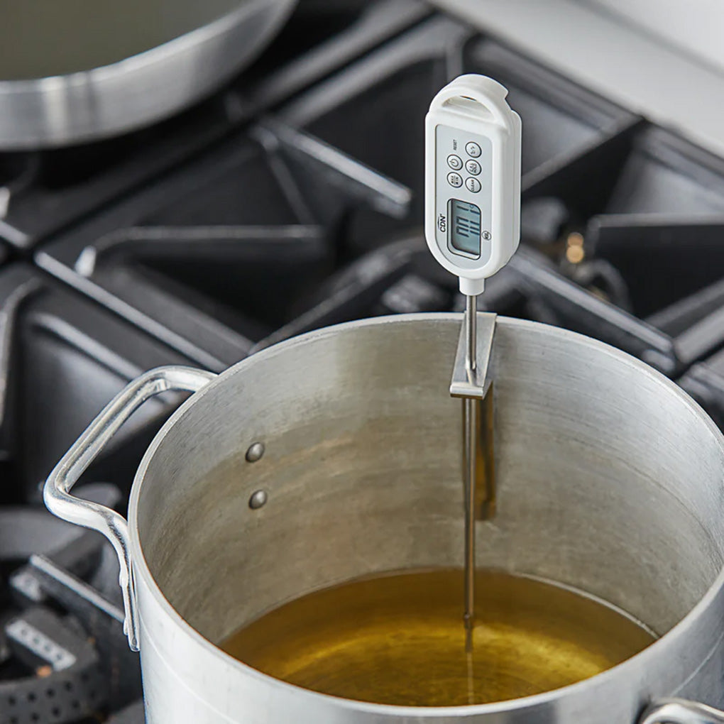 Digital thermometer clipped to the rim of a large pot filled with oil on a stovetop, showing the long probe submerged and the display facing forward.