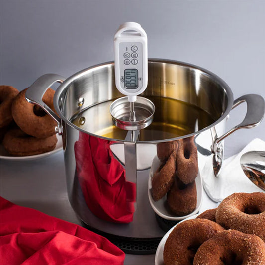 Digital thermometer clipped to the side of a stainless steel pot of hot oil, with donuts on plates nearby and the temperature display visible.