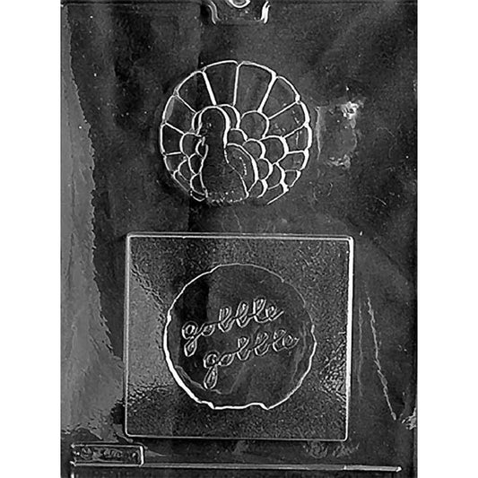 Clear “Gobble Gobble” Turkey Disk & Tray Chocolate Mold photographed against a black background with a detailed turkey disk cavity and coordinating tray cavity with raised Gobble Gobble lettering.