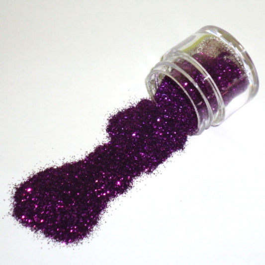 Small clear jar tipped on its side spilling grape purple glitter dust onto a white surface, with fine sparkling particles visible throughout.