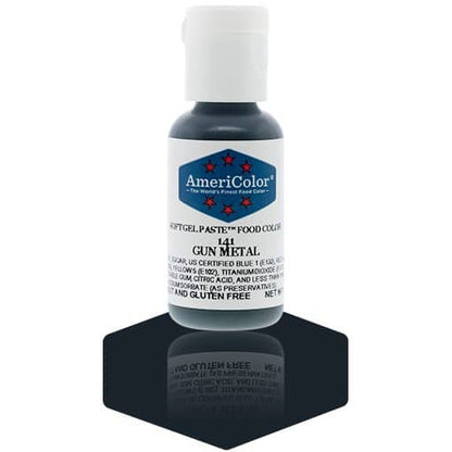 A bottle of AmeriColor Gun Metal soft gel paste food coloring with a white cap, blue label, and red stars, positioned on a dark gunmetal-gray reflective surface with the bottle’s reflection clearly visible.