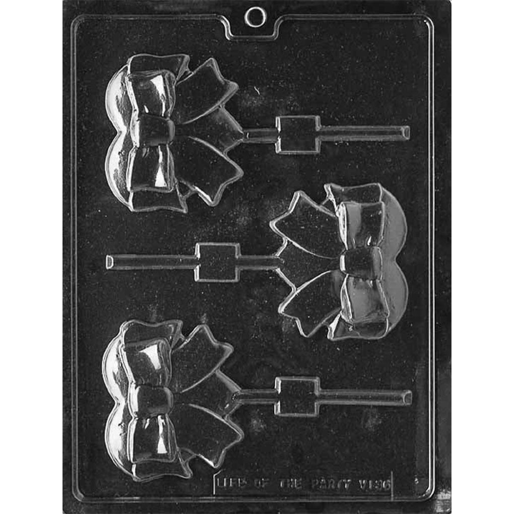 Clear plastic chocolate lollipop mold with three cavities shaped like a heart wrapped in a large raised bow and ribbon tails, each connected to a stick channel on a dark background.