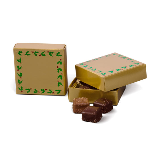 Gold square two-piece candy box with a green holly and red berry border, shown closed and open with assorted chocolates beside it on a white background.