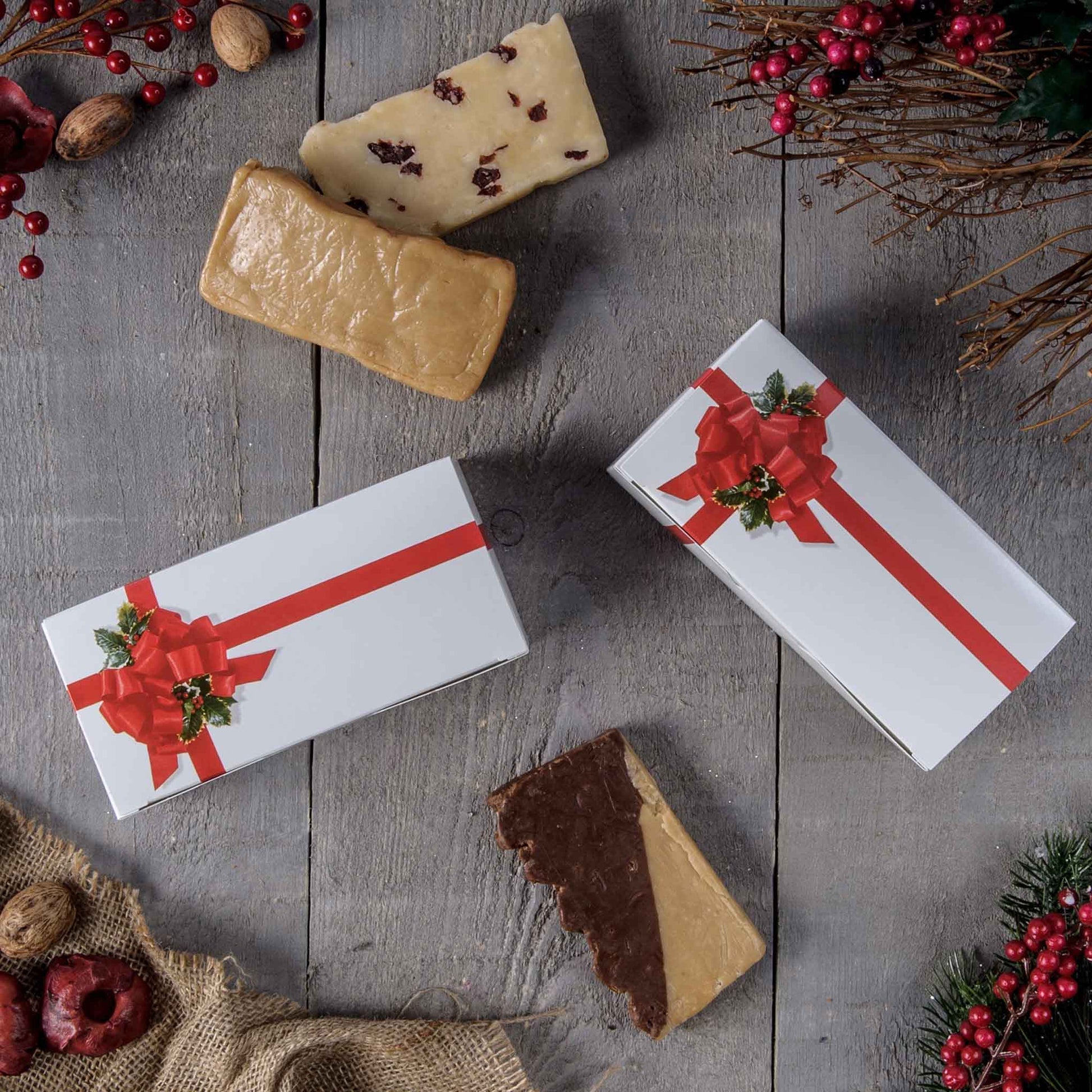 Holiday flat-lay with white ribbon-and-holly candy boxes on a rustic wood surface, surrounded by festive berries, greenery, and assorted candy pieces.