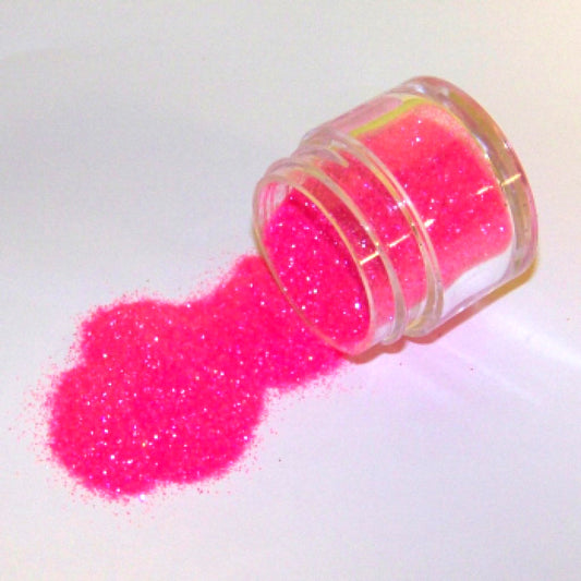Small clear jar tipped on its side spilling bright hot pink glitter dust onto a white surface, with fine sparkling particles visible throughout.