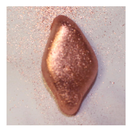 Close-up of a rose gold metallic shimmer swatch on a white surface with scattered sparkling dust around it, showing a warm coppery pink sheen.