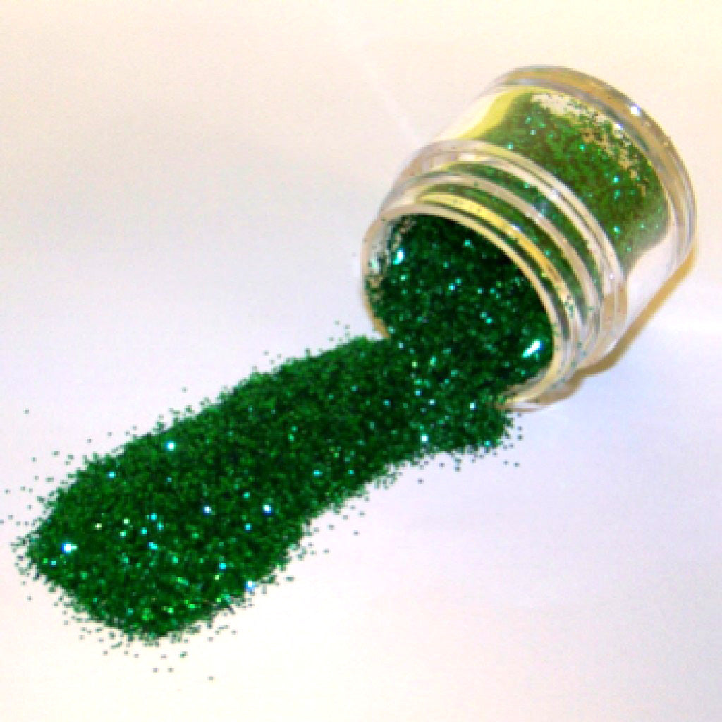 Small clear jar tipped on its side spilling kelly green glitter dust onto a white surface, showing dense sparkling green particles.