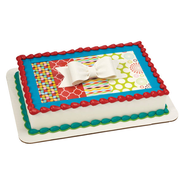 Rectangular birthday cake with colorful patterns and a large white gum paste bow as the centerpiece topper.