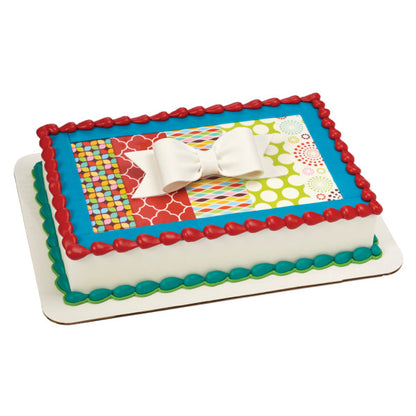 Rectangular birthday cake with colorful patterns and a large white gum paste bow as the centerpiece topper.