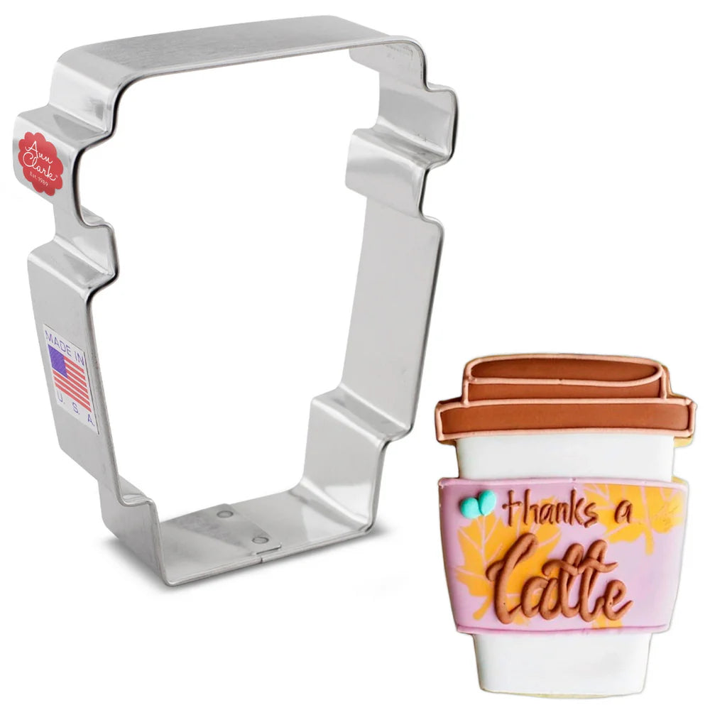 Metal latte cup cookie cutter alongside a decorated coffee cup cookie with 'thanks a latte' written on a pink and orange sleeve.