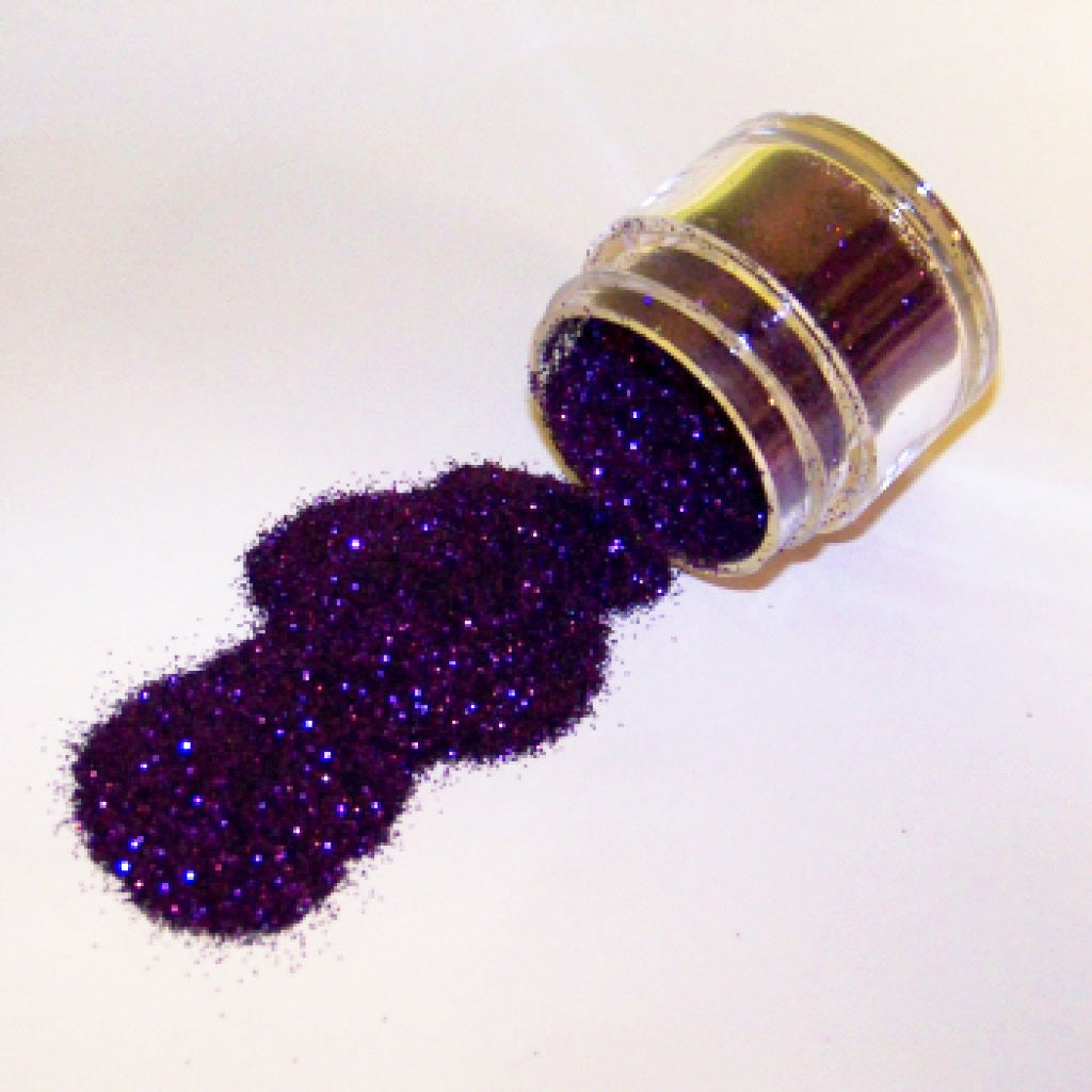 Small clear jar tipped on its side spilling deep lilac-purple glitter dust onto a white surface, showing fine sparkling particles for cake decorating.