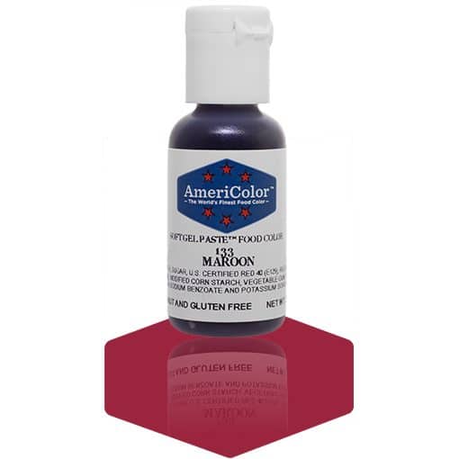 A bottle of AmeriColor Maroon soft gel paste food coloring with a white cap, blue label, and red stars, standing on a deep maroon reflective surface with the bottle’s reflection visible.