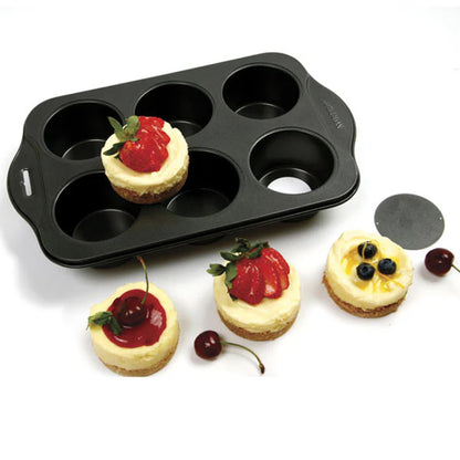 Six-cavity mini cheesecake pan on a white surface with four finished mini cheesecakes topped with strawberries, cherries, lemon slices, and blueberries; a round removable bottom disk sits nearby.