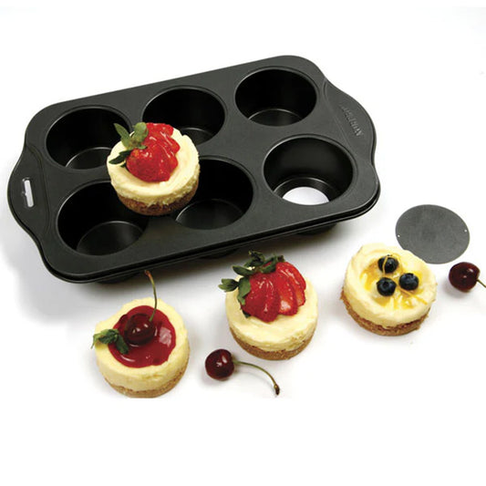 Six-cavity mini cheesecake pan on a white surface with four finished mini cheesecakes topped with strawberries, cherries, lemon slices, and blueberries; a round removable bottom disk sits nearby.