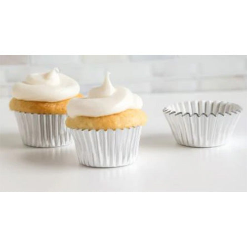 Two mini cupcakes in silver foil baking cups with white frosting, beside an empty silver foil cup on a light surface.