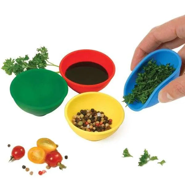 Four flexible silicone prep bowls in red, yellow, blue, and green, each containing ingredients like chopped herbs, oil, and whole spices.