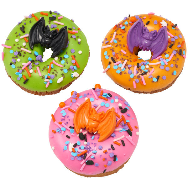 Three Halloween donuts with pink, green, and orange icing, topped with Monster Mash Deluxe Sprinkle Blend and plastic bat toppers.