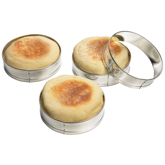 Four shiny round metal muffin rings on a white background, with three rings holding golden-brown English muffins and one empty ring leaning against them.