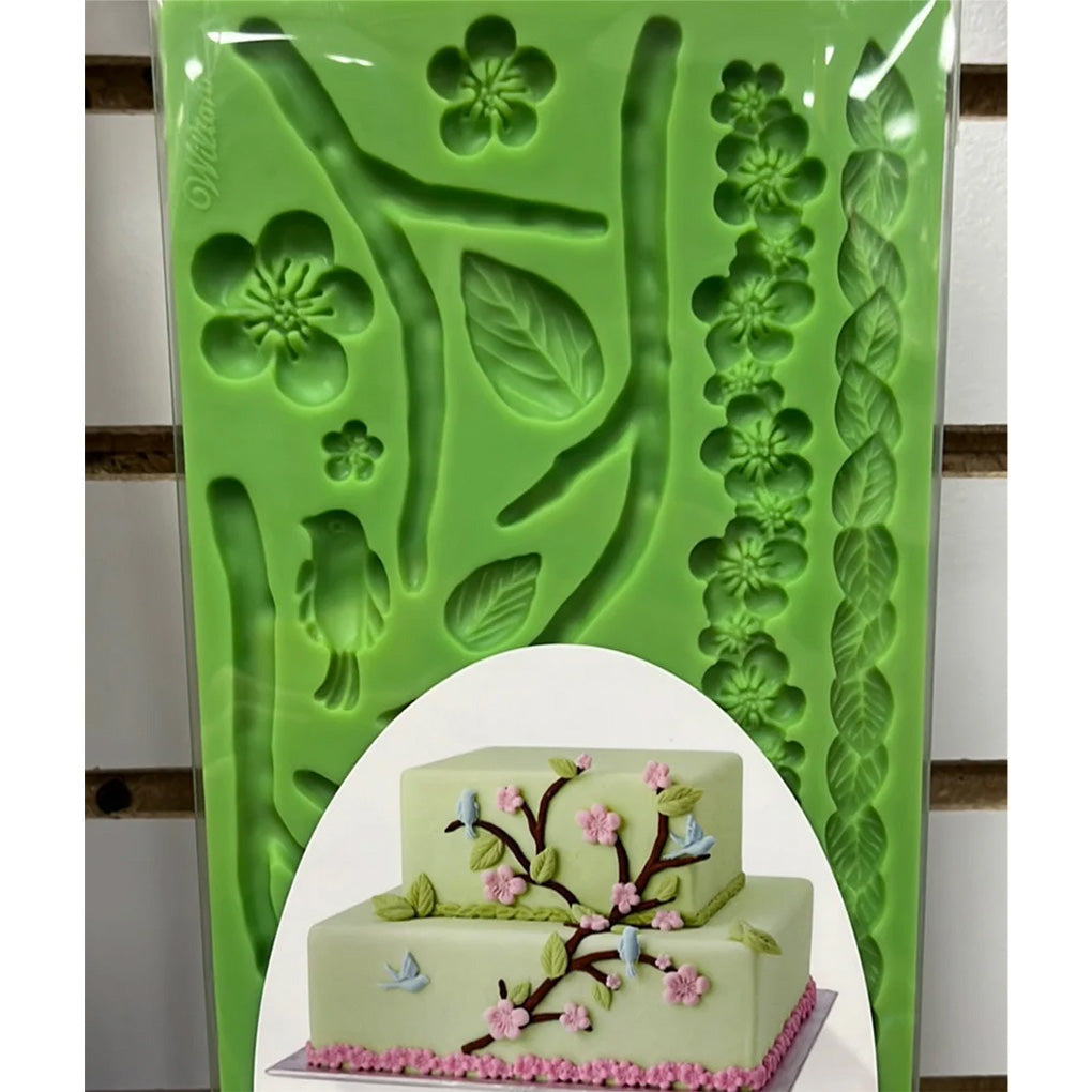 Green silicone fondant and gum paste mold with cavities for curved branches, multiple leaves, five-petal flowers, a small bird shape, and long leaf and flower border strips; shown with a sample decorated cake image on the packaging.
