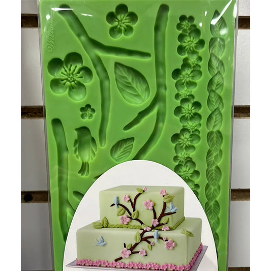 Green silicone fondant and gum paste mold with cavities for curved branches, multiple leaves, five-petal flowers, a small bird shape, and long leaf and flower border strips; shown with a sample decorated cake image on the packaging.