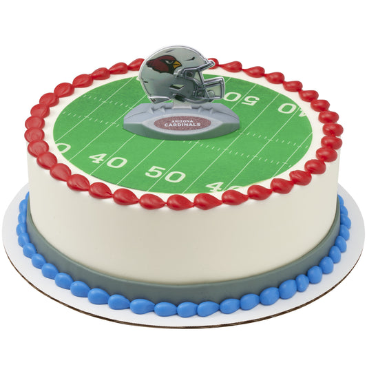 A round cake decorated to look like a football field, with a green yard-line surface, red piped border, blue base trim, and a small Arizona Cardinals helmet topper on top.