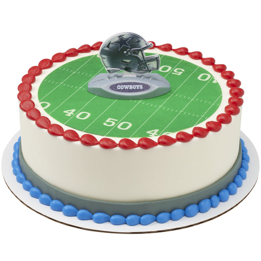 A round cake decorated with a green football field design, red and blue piped borders, and a Dallas Cowboys helmet topper on top.