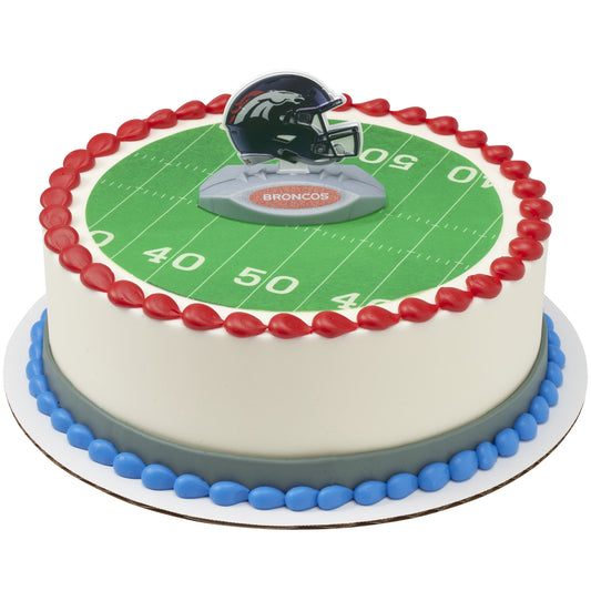 A round cake decorated with a green football field print, red piped border, blue base detail, and a Broncos helmet topper on top.